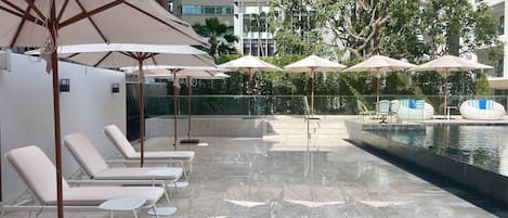 A poolside area with lounge chairs and umbrellas, a water feature, and a view of a modern building.