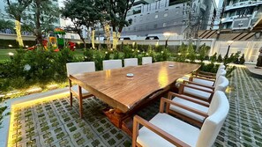 An outdoor dining area with a wooden table and white cushioned chairs. - Floral Court Hotel and Residence (Bangkok)