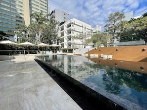 A swimming pool with a wooden deck and lounge chairs, surrounded by buildings and trees. - Floral Court Hotel and Residence (Bangkok)