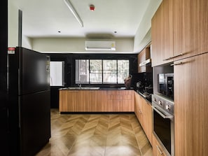 A modern kitchen with wooden cabinets, a black refrigerator, a stove, and a window providing natural light. - Floral Court Hotel and Residence (Bangkok)
