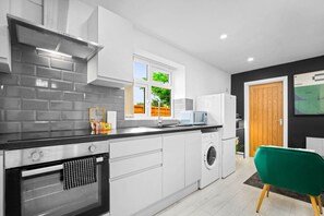 A modern kitchen with white cabinets, a black countertop, a built-in oven, a washing machine, and a window with a view of greenery outside.