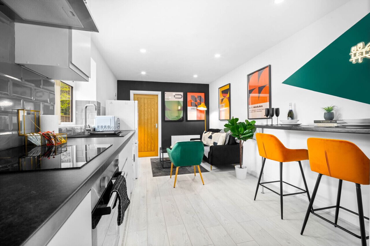 A modern kitchen with a black countertop, white cabinets, and a wooden door. There are two chairs, one green and one orange, and a small table with a plant.