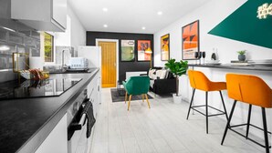 Een moderne keuken met een zwart aanrecht, witte kastjes en een houten deur. Er staan twee stoelen, een groene en een oranje, en een kleine tafel met een plantje.