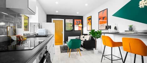 A modern kitchen with a black countertop, white cabinets, and a wooden door. There are two chairs, one green and one orange, and a small table with a plant.