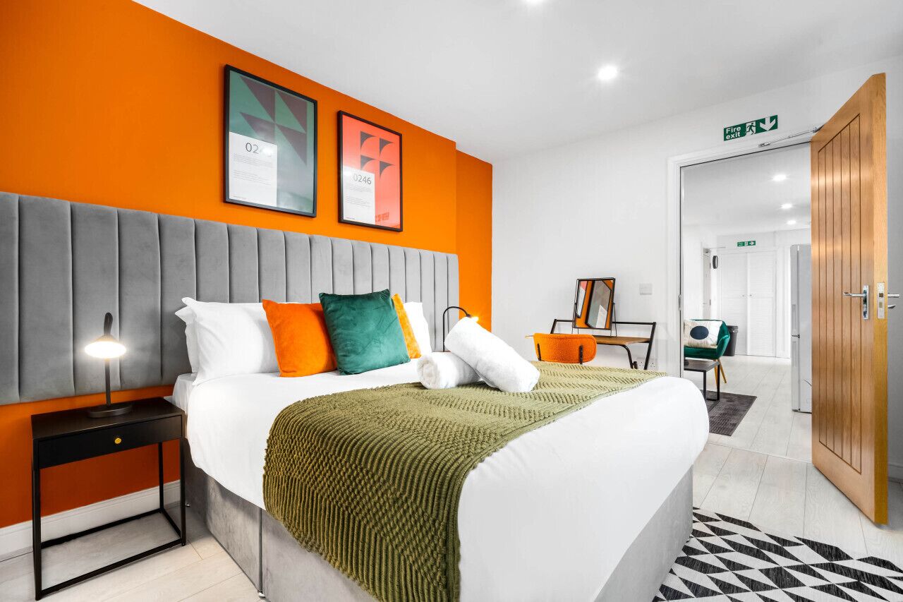A hotel room with a bed, a nightstand, a lamp, a green throw blanket, and a framed picture on the wall.
