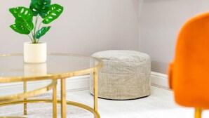 Un coin de pièce avec une table ronde en verre, un pouf beige et une plante en pot.