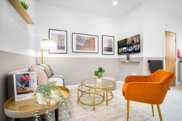 Una sala moderna con sofá, un sillón naranja, una mesa de centro redonda de vidrio y cuadros en la pared.