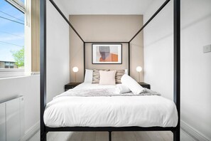 A modern bedroom with a bed, bedside lamps, a framed artwork, and a window.