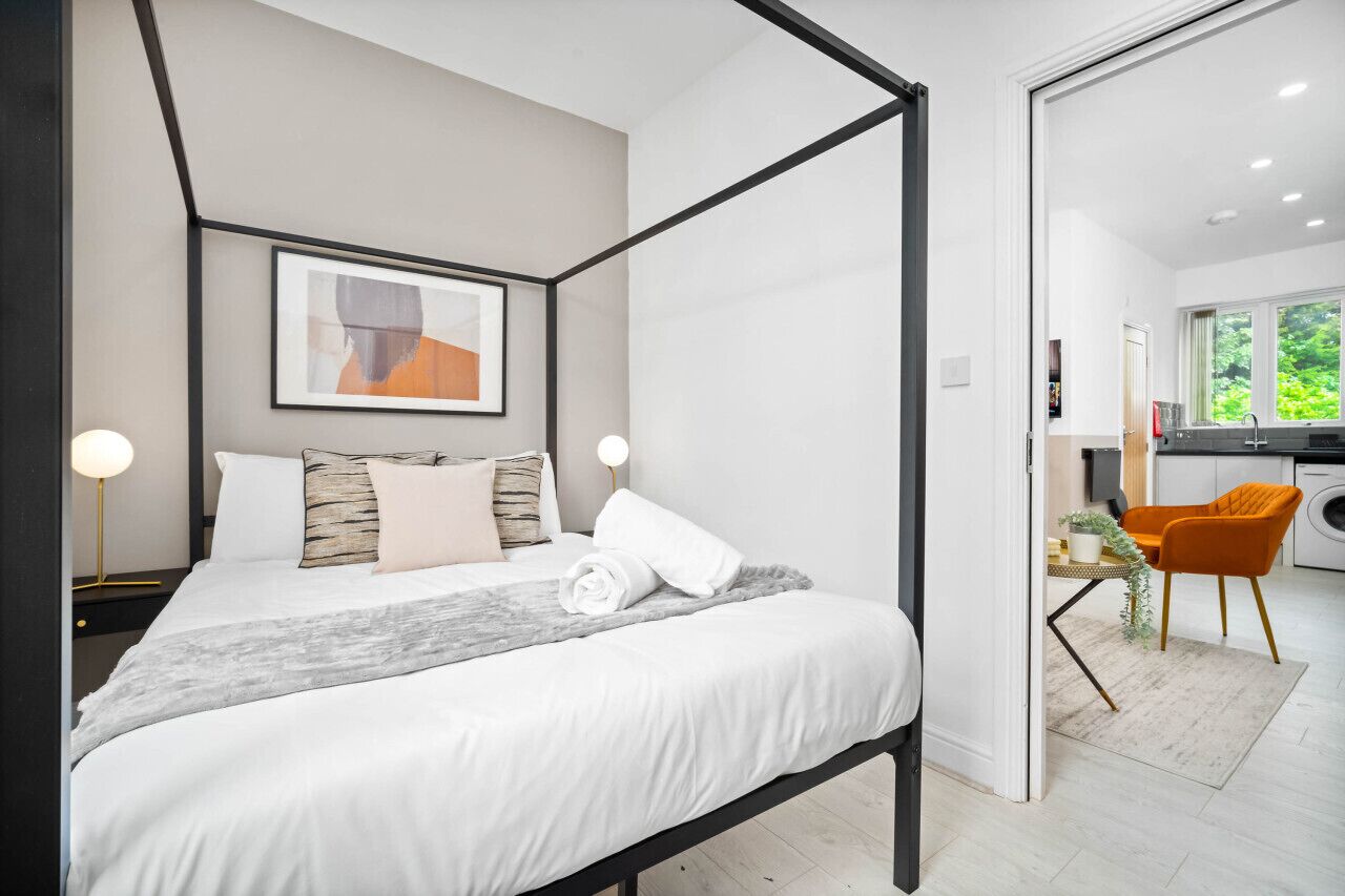 Une chambre moderne avec un lit, des lampes de chevet, une œuvre d’art encadrée et une vue sur la cuisine.