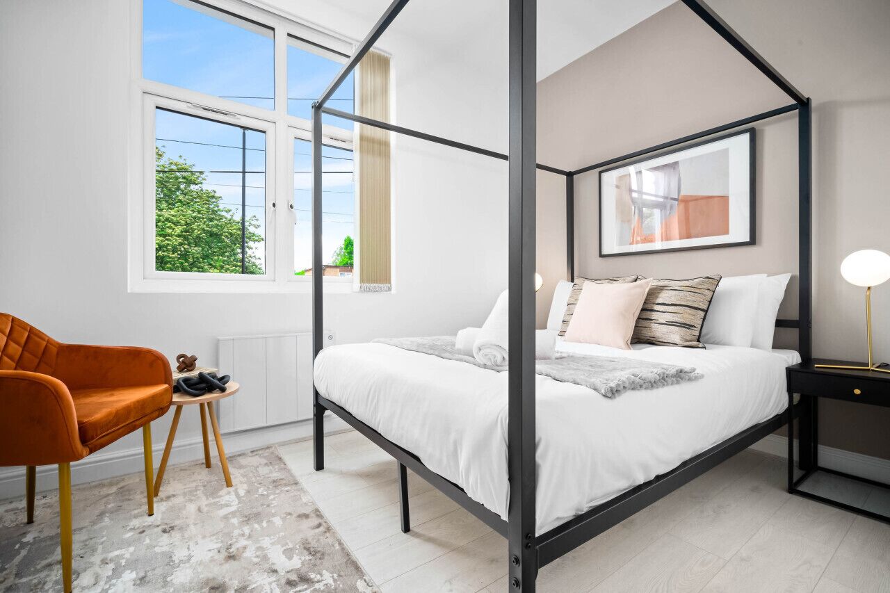 A modern bedroom with a four-poster bed, a small orange chair, a bedside table with a lamp, and a window with a view of greenery.