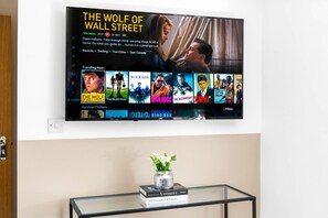 Un téléviseur à écran plat est fixé au mur et affiche une affiche du film « Le Loup de Wall Street », ainsi qu’une sélection d’autres films. En dessous du téléviseur, on trouve une table en verre sur laquelle sont posés un vase de fleurs et un livre intitulé « Lieux délicieux ».