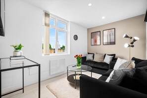 A modern living room with a black sofa, a glass coffee table, and a white radiator. There is a window with a view of buildings outside. - Luxury 2 Bedroom Apartment  - WiFi - Smart TV (Birmingham)