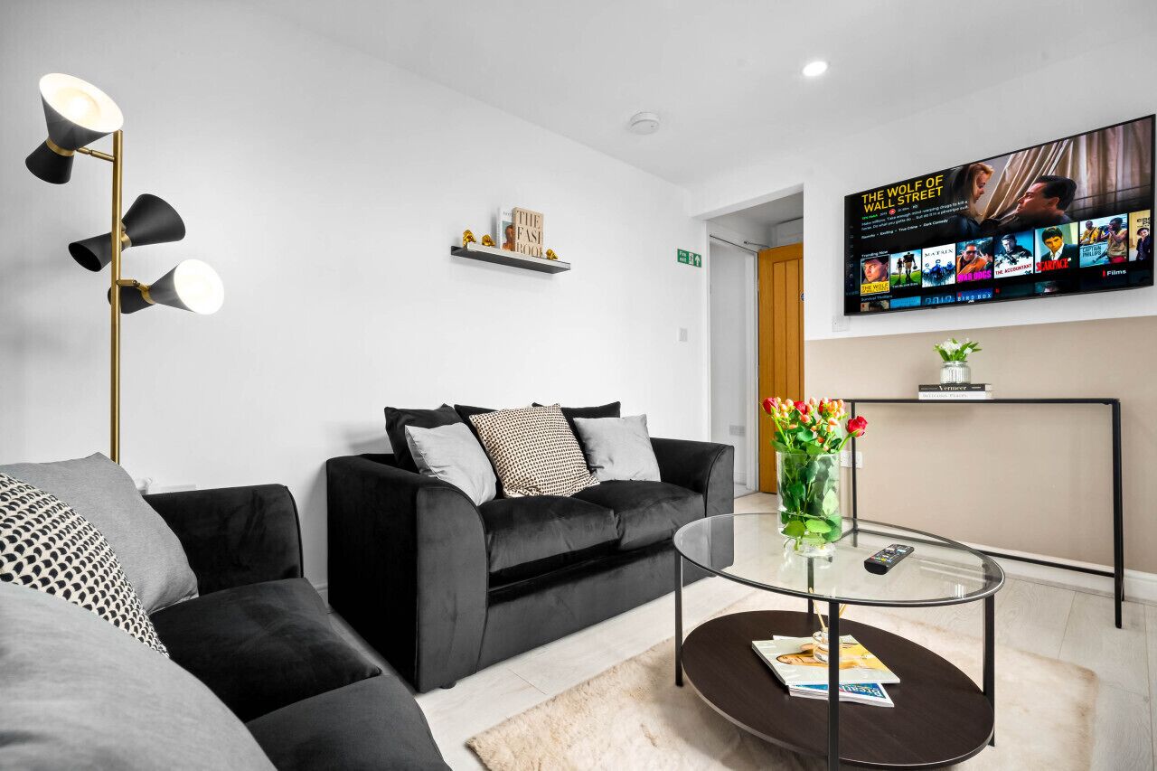 A modern hotel lobby with a black sofa, a glass coffee table, a floor lamp, and a TV displaying movie posters.