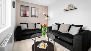 En moderne stue med to sorte sofaer, et glasbord, en vase med blomster og et bogreol med bøger og pyngeting.