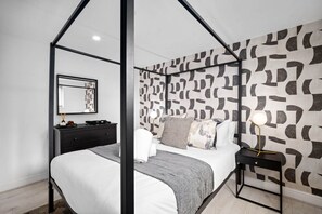 A modern bedroom with a four-poster bed, a black nightstand with a lamp, a dresser, and a patterned wall.