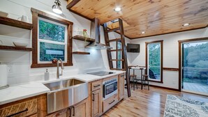 A modern kitchen with wooden cabinetry, a stainless steel sink, and a built-in oven. There is a wooden ladder leaning against the wall, and a window overlooking a deck. - Cozy Couples Treehouse w Hot Tub, Fire Pit, Pet Friendly, Lake Access (Morganton)