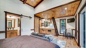 A bedroom with a large bed, a wooden ceiling, a kitchen area with a window, and a TV mounted on the wall. - Cozy Couples Treehouse w Hot Tub, Fire Pit, Pet Friendly, Lake Access (Morganton)