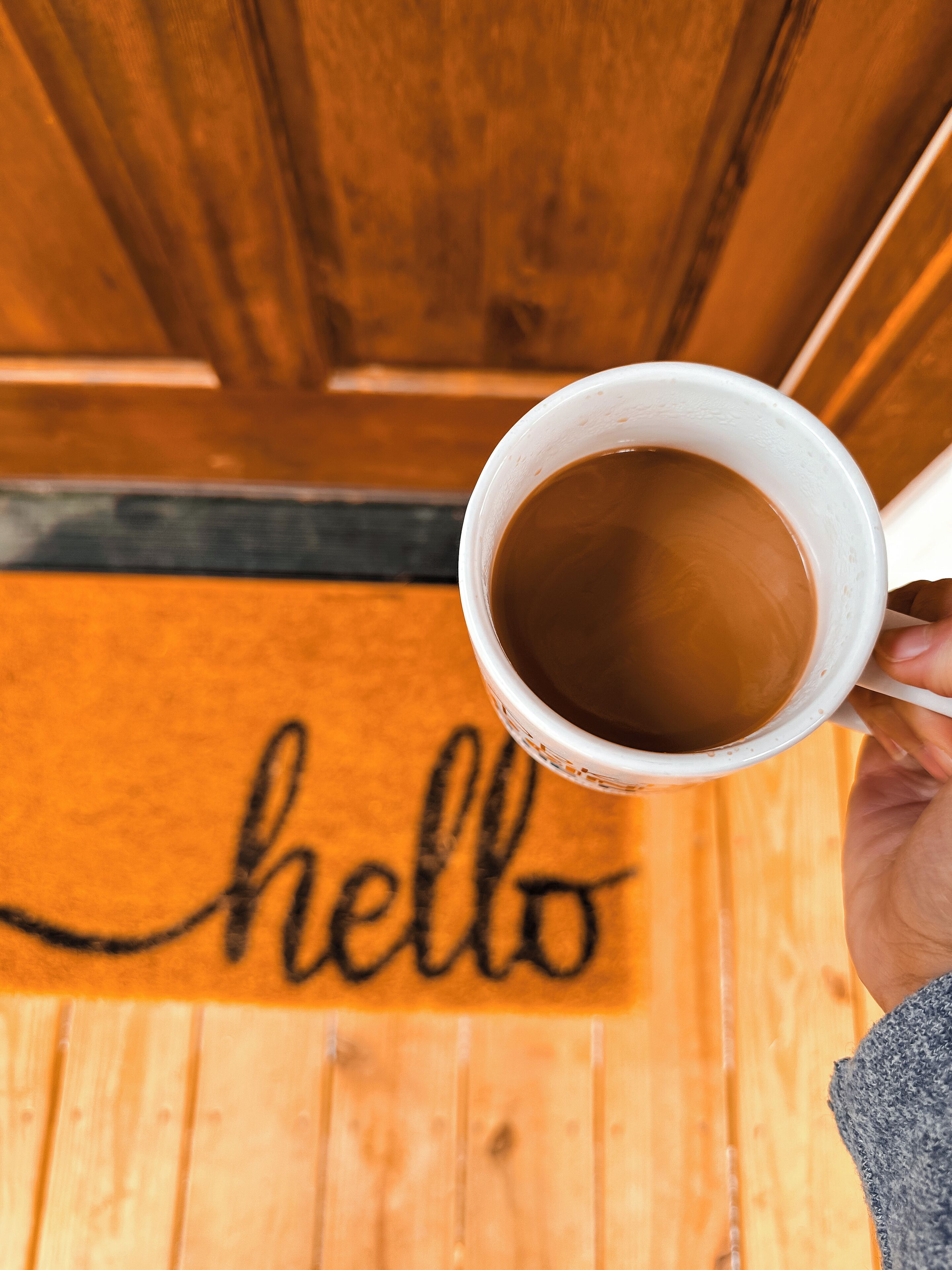 A hand holding a white mug with a brown liquid, likely coffee, in front of a wooden door with a "hello" mat.