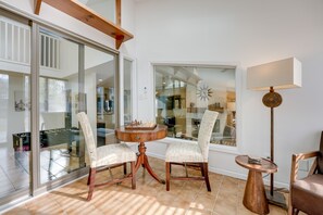 Une pièce dotée d’une porte vitrée donnant sur un balcon, une table ronde en bois avec un jeu d’échecs, deux fauteuils rembourrés, un lampadaire et une vue sur le paysage urbain.