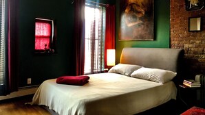 Une chambre d’hôtel avec un grand lit, un rideau rouge, une fenêtre avec des stores, un tableau au mur et un mur en briques.