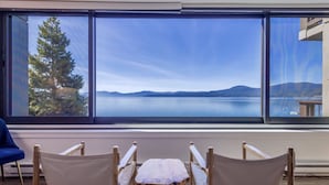 Une pièce dotée d’une grande fenêtre donnant sur un lac et des montagnes, une table en bois avec des chaises et un fauteuil bleu.
