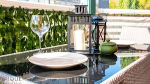Une table en verre sur laquelle sont posés une assiette blanche, un verre à vin, une bougie et une théière verte, le tout sur un balcon doté d’une rambarde en métal et dont l’arrière-plan est verdoyant.