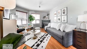 Un salon moderne avec un canapé gris, une table basse et une cheminée. Des tableaux encadrés ornent le mur, et une plante est posée sur le rebord de la fenêtre.