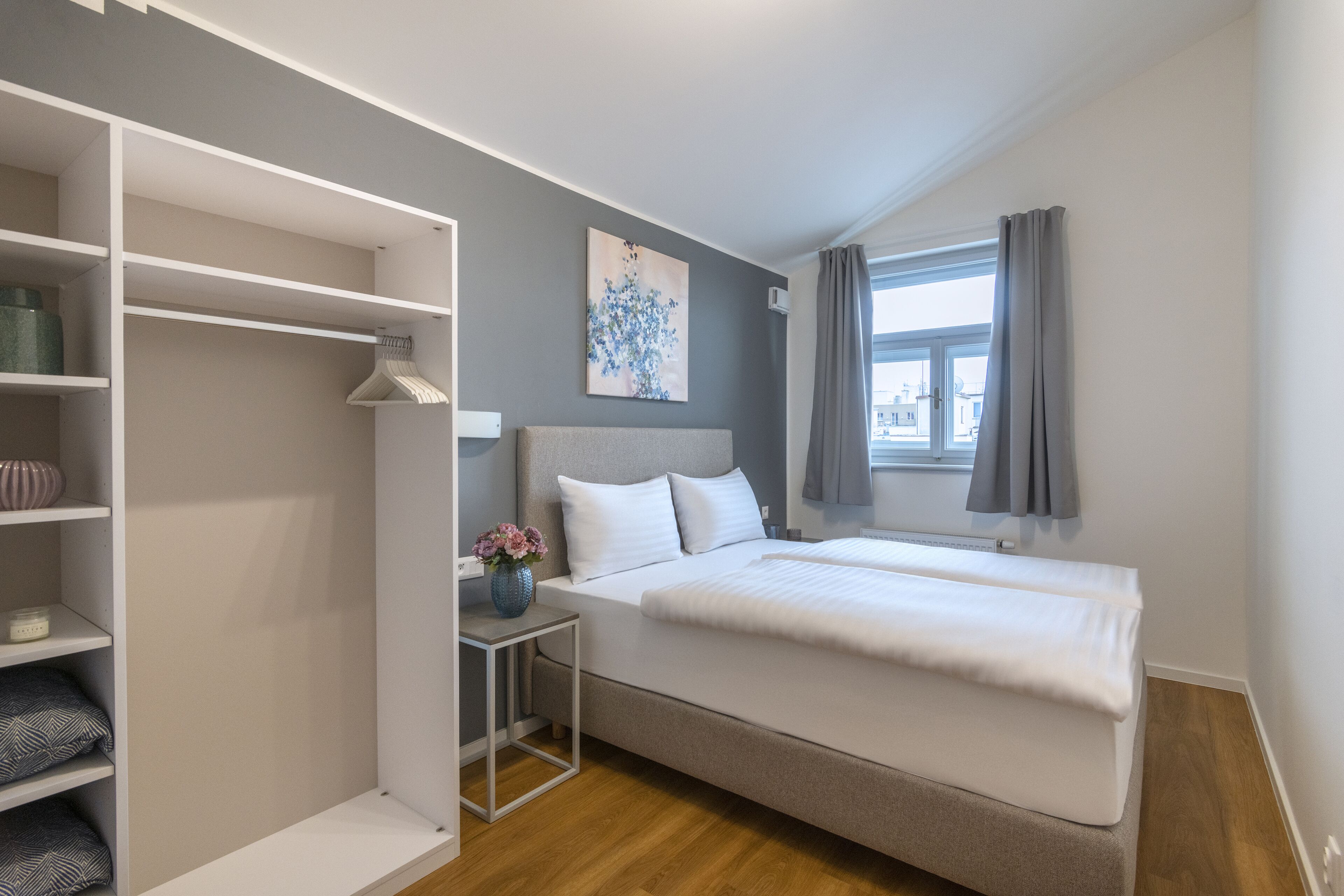 Ein Schlafzimmer mit einem Bett, einem Nachttisch mit einer Vase, einem Kleiderschrank und einem Fenster mit Vorhängen.