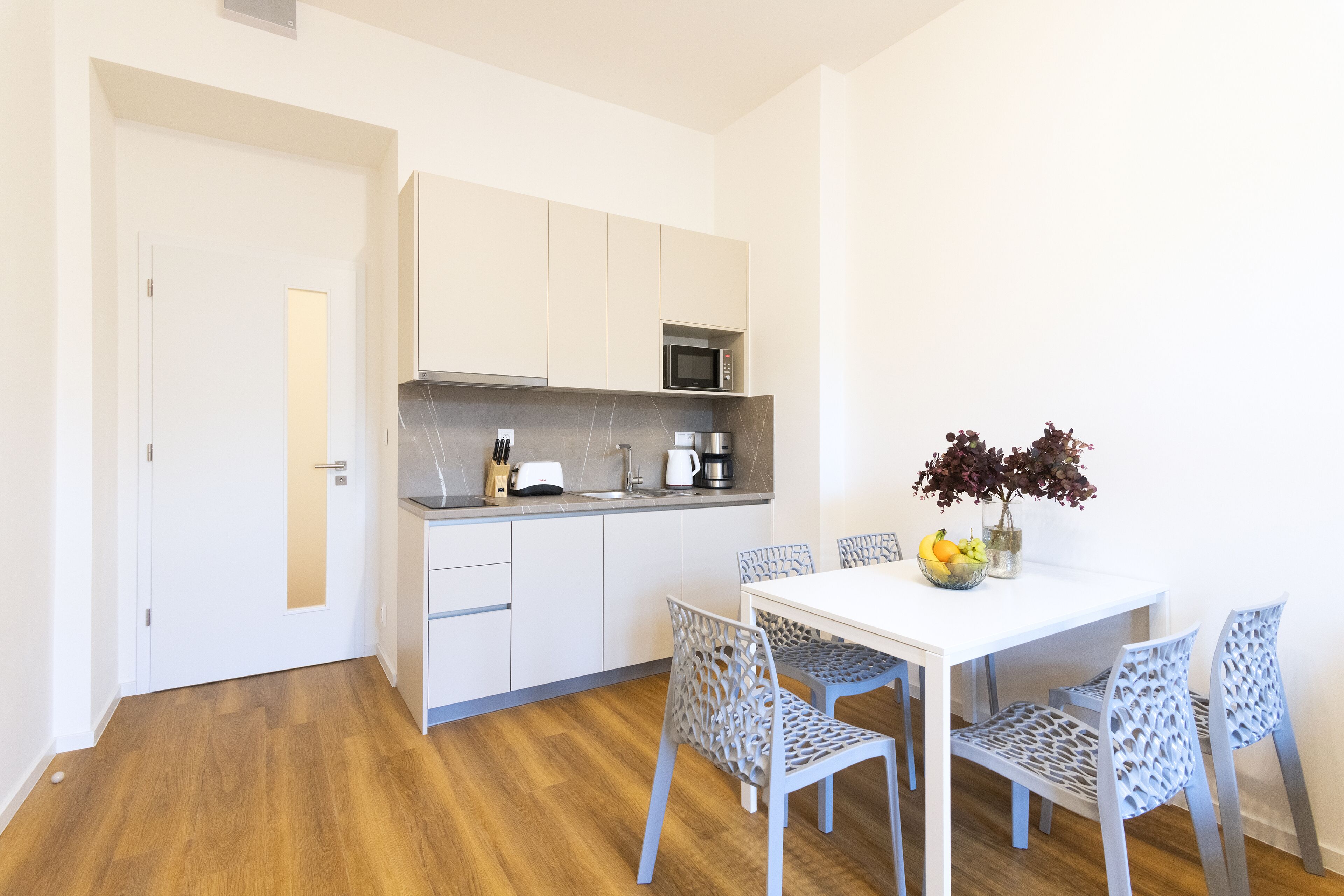 Una cocina moderna con armarios blancos, microondas y cafetera. Hay una mesa de comedor con cuatro sillas y un florero con flores.