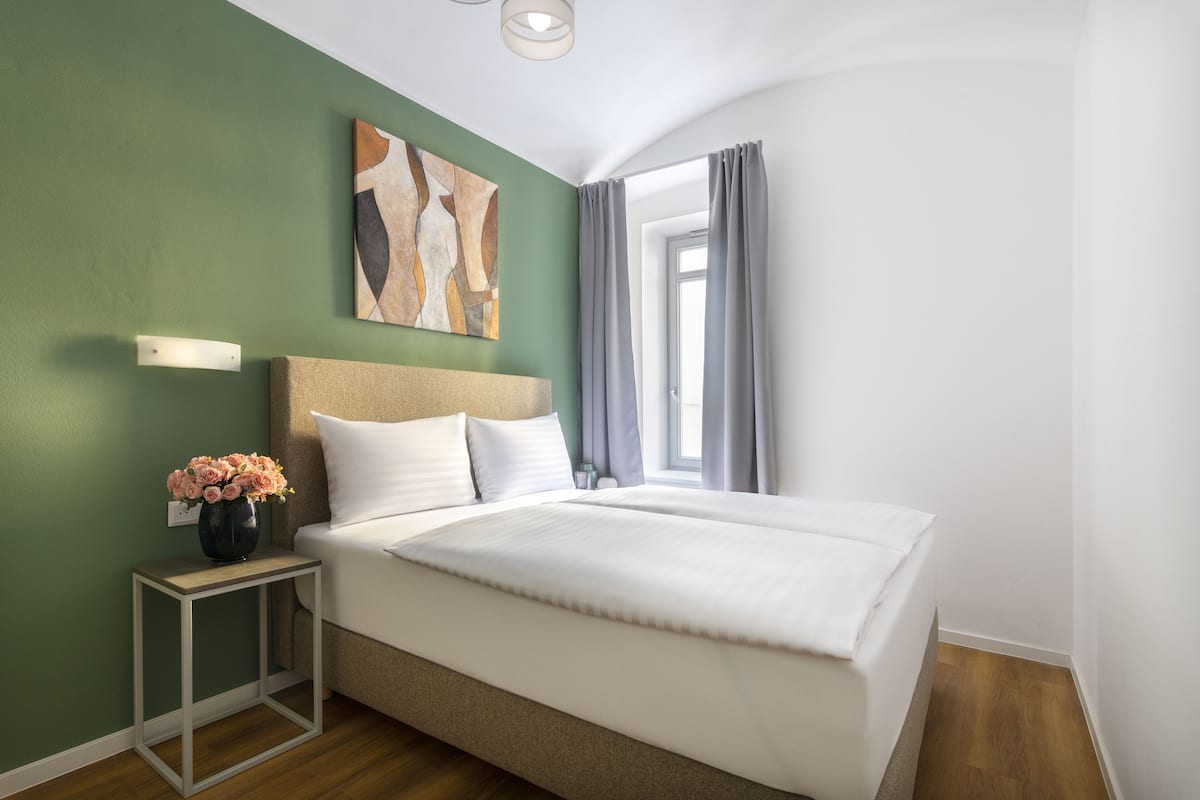 Una habitación de hotel moderna con una cama grande, una mesita de noche con un jarrón de flores, una obra de arte en la pared y una ventana con cortinas.