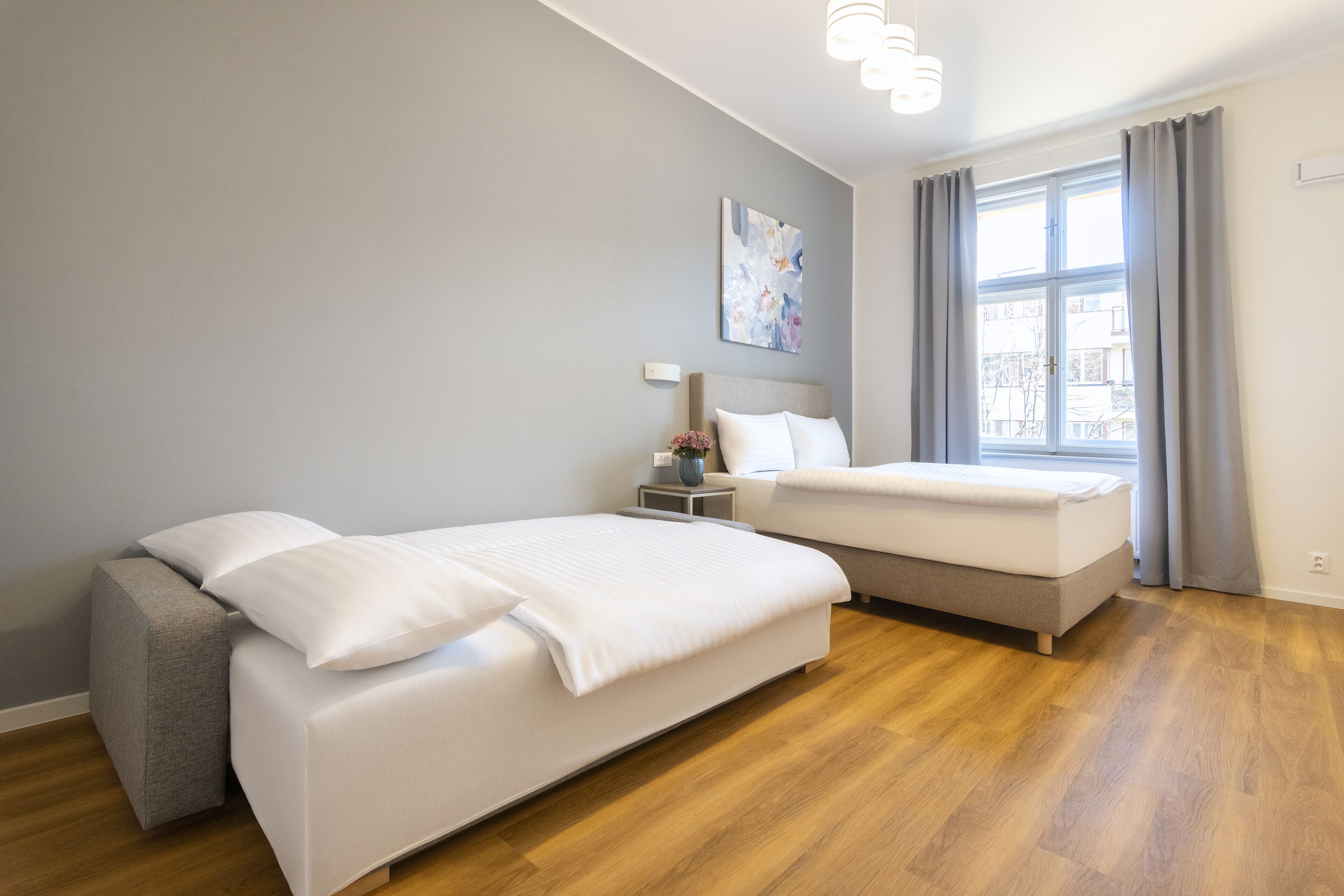 Una moderna camera d'hotel con due letti, un'ampia finestra con tende, un comodino con un vaso e un quadro appeso al muro.