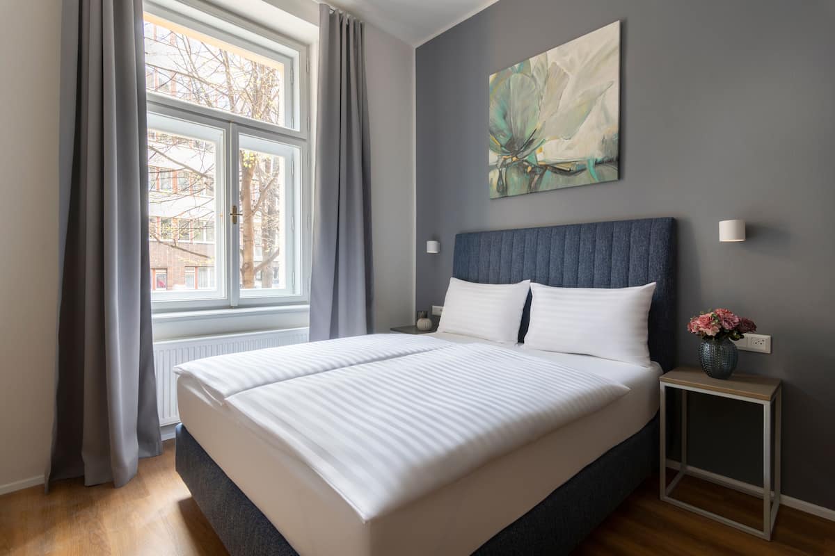 Una cama bien tendida en una habitación con un ventanal grande, una pared gris y un cuadro sobre la cama.