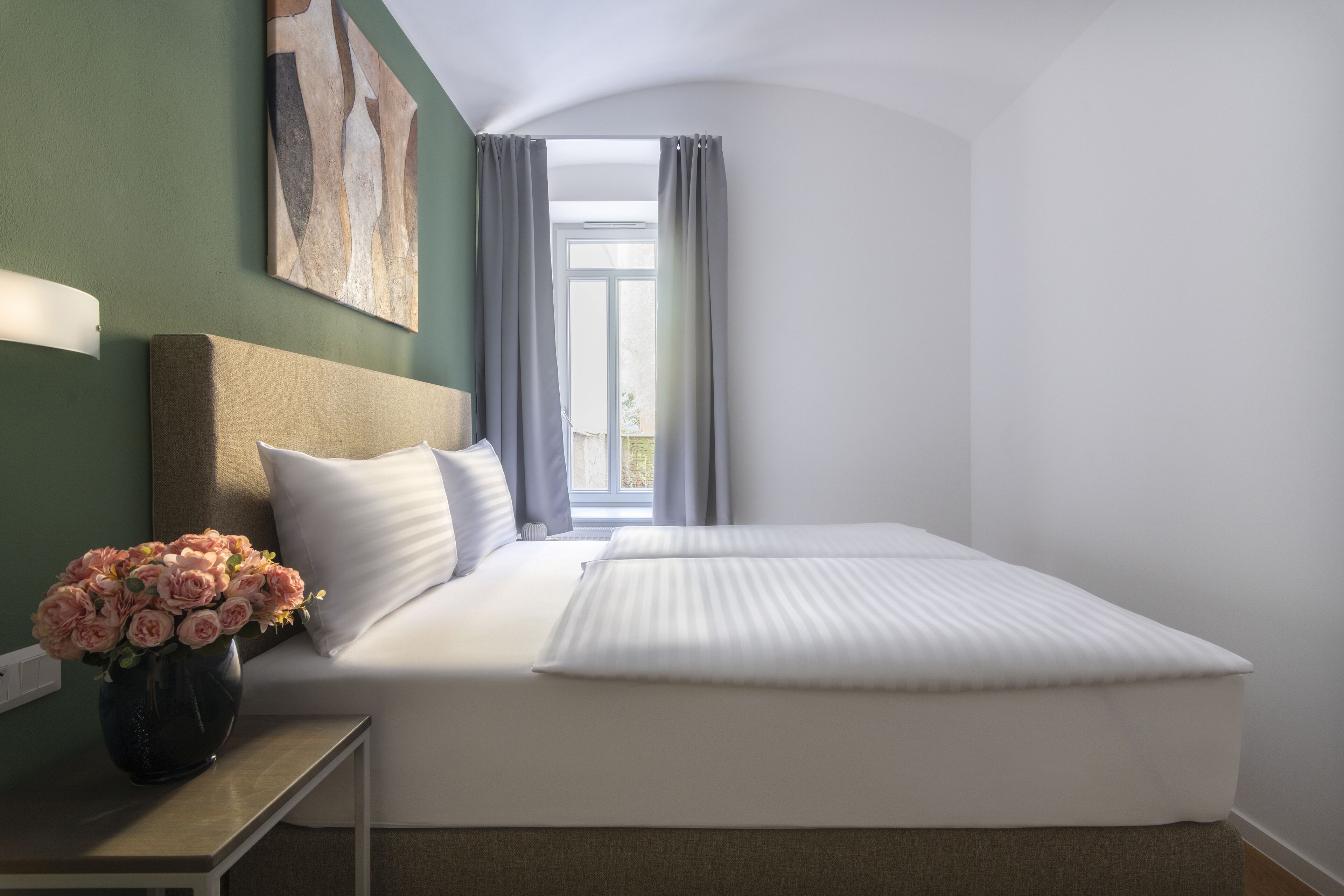 Una cama bien hecha con sábanas blancas, un jarrón de flores rosas sobre una mesita de noche y un cuadro en la pared.