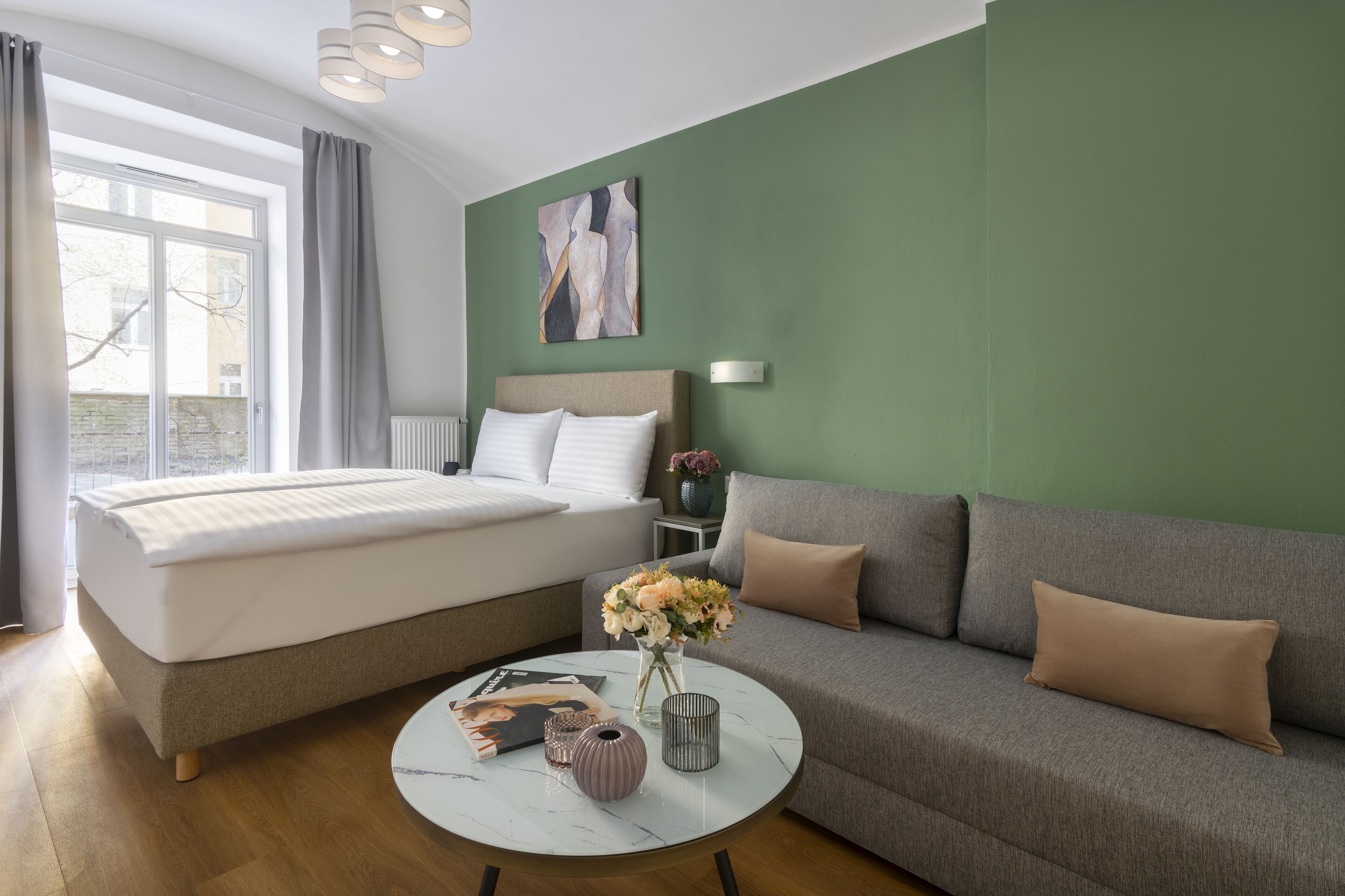 Moderni hotellihuone, jossa on sänky, sohva, koriste-esineillä varustettu pyöreä sohvapöytä ja näkymä ulos.
