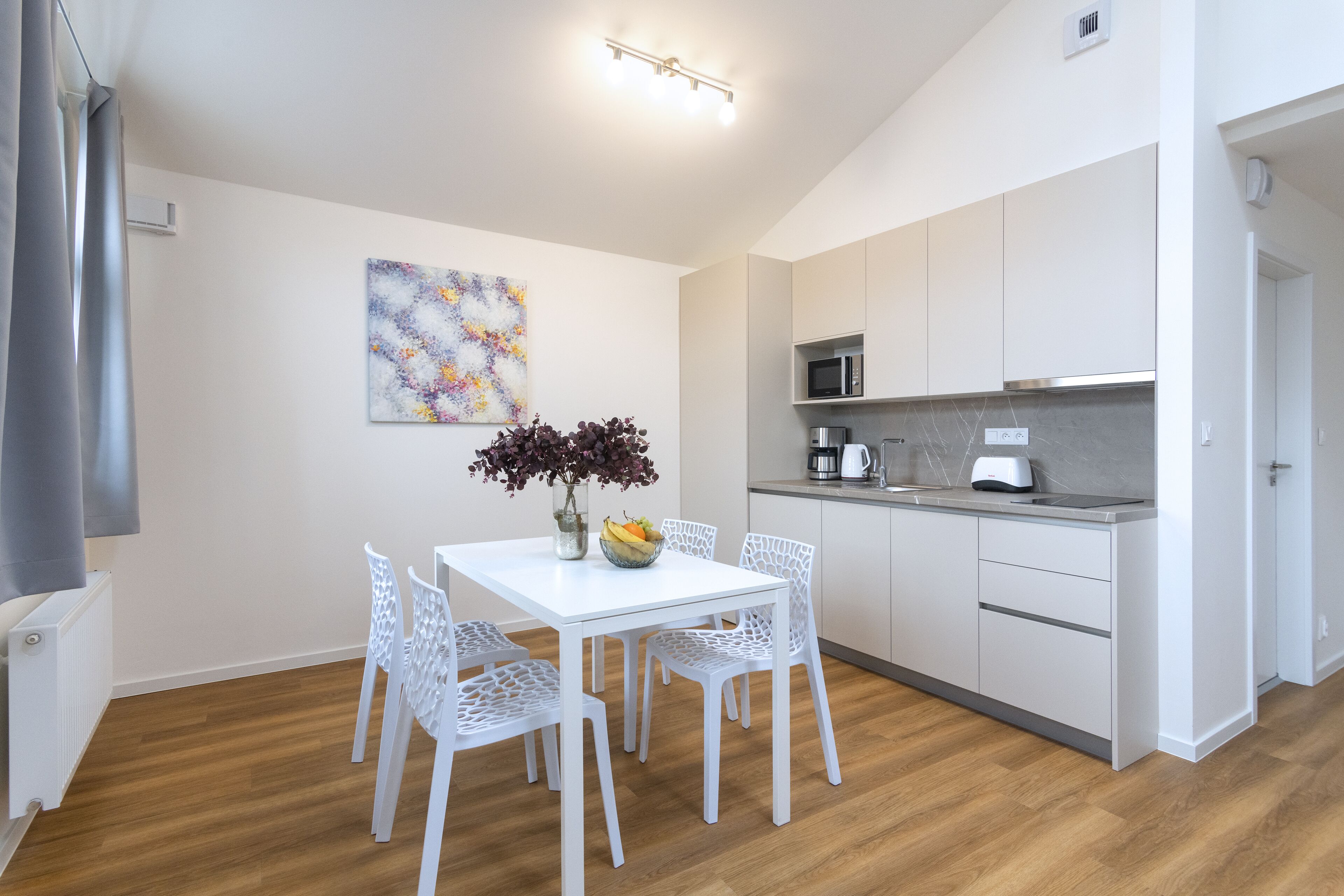A modern kitchen with white cabinets, a microwave, and a coffee maker. There is a dining table with white chairs and a vase with flowers.