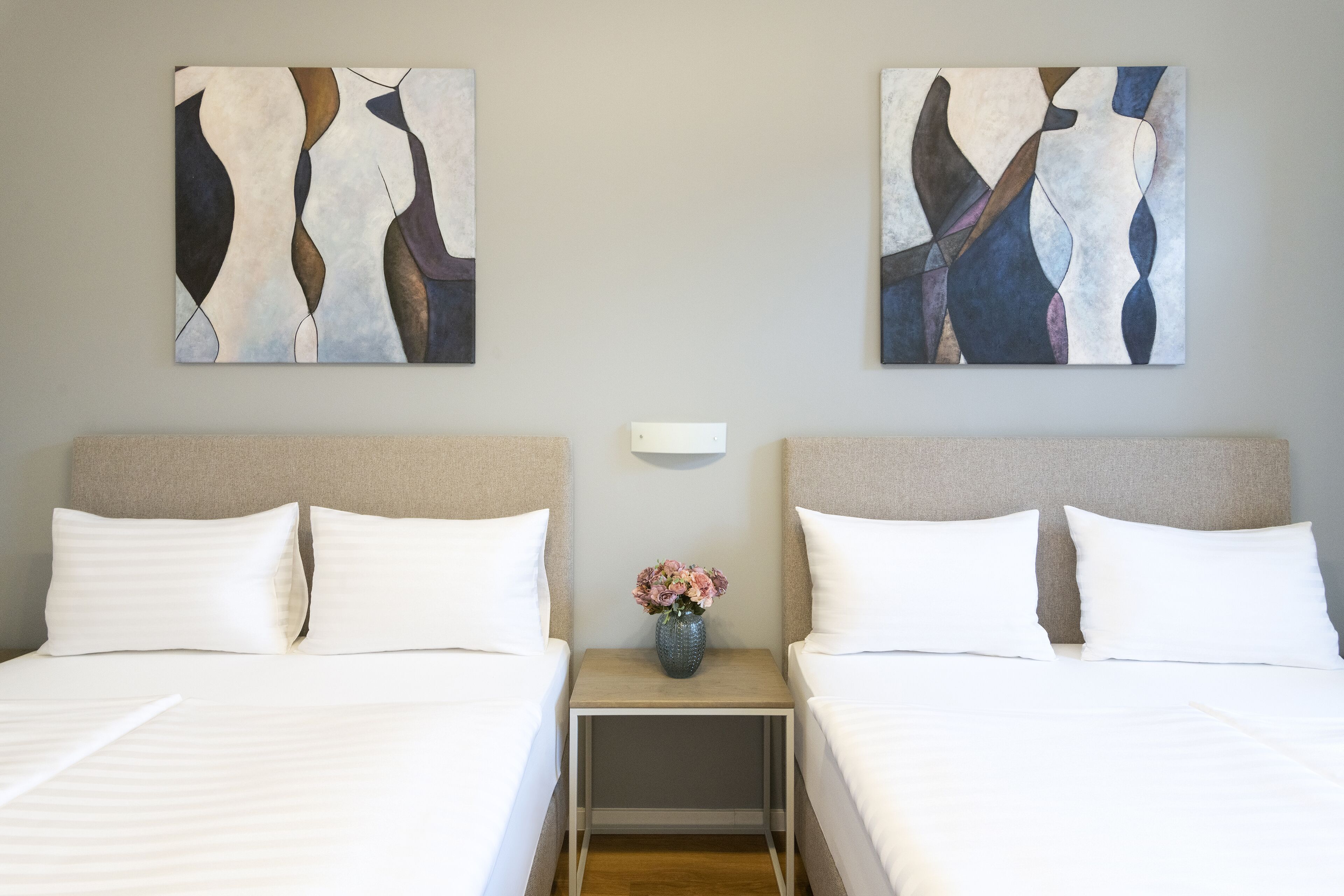 A hotel room with two beds, a nightstand with a vase of flowers, and abstract art on the walls.
