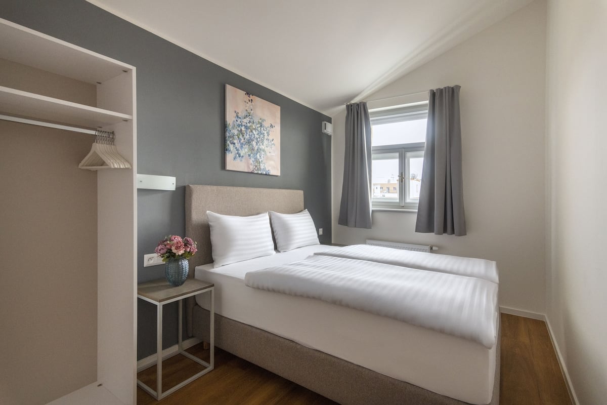 Un dormitorio con dos camas, una mesita de noche con un florero, un perchero y una ventana con cortinas.