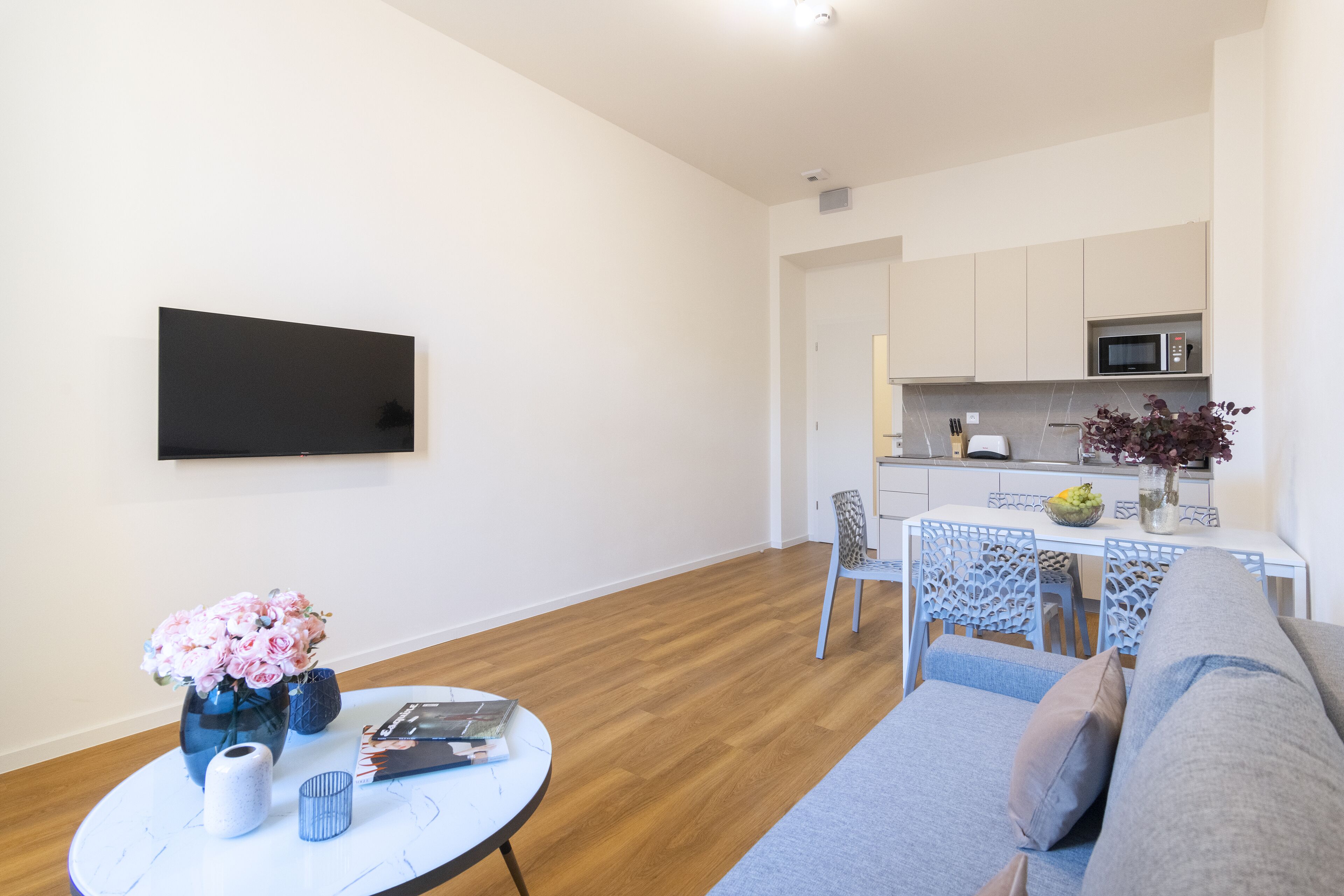 Uma sala de estar moderna com um sofá, uma mesa redonda com um vaso de flores, uma televisão de tela plana na parede e uma área de cozinha com micro-ondas e uma mesa de jantar.
