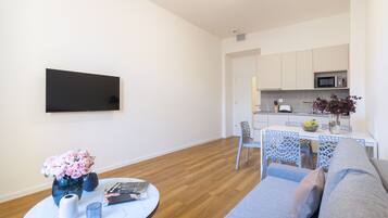 Uma sala de estar moderna com um sofá, uma mesa redonda com um vaso de flores, uma televisão de tela plana na parede e uma área de cozinha com micro-ondas e uma mesa de jantar.