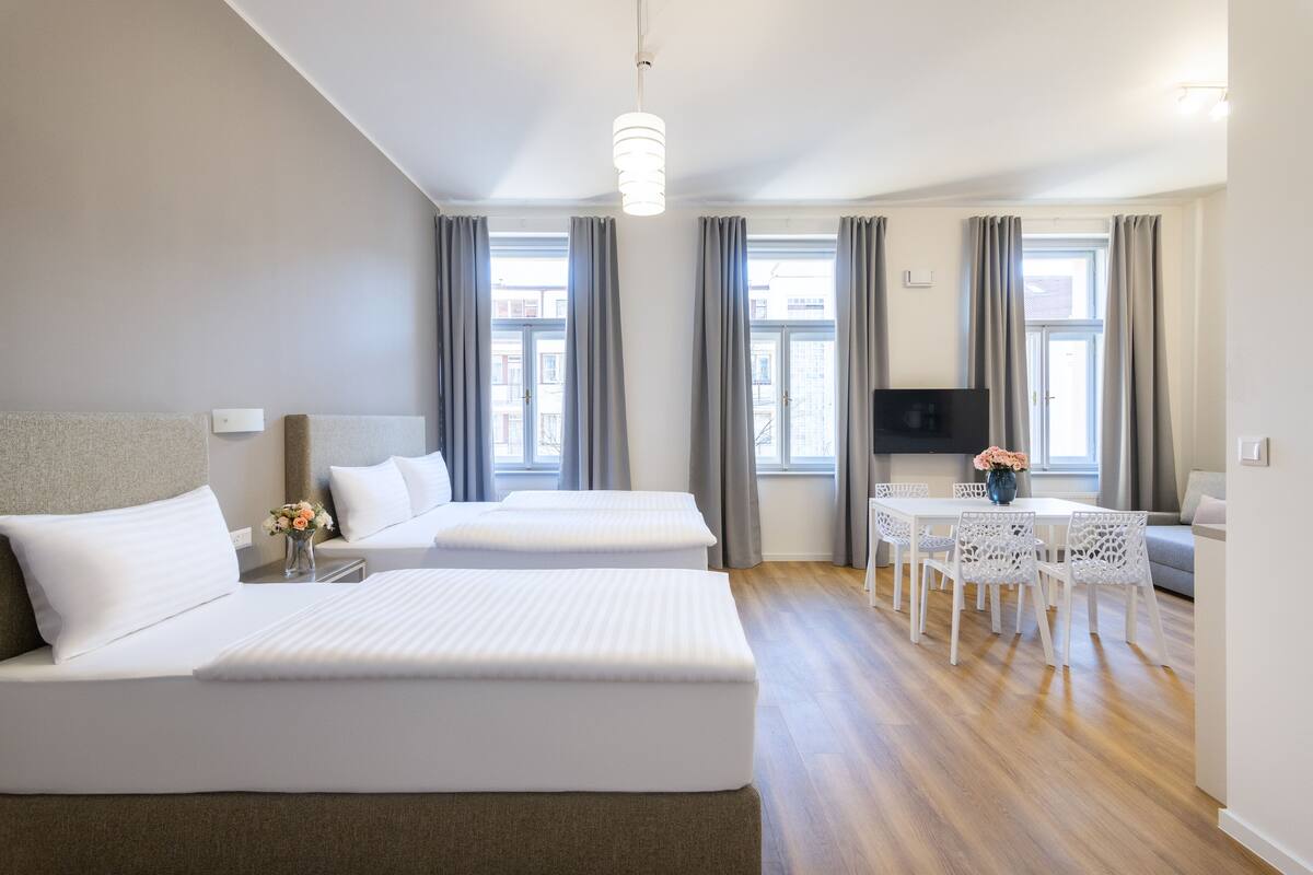 Habitación de hotel con dos camas, una mesa de comedor con sillas, un televisor y una ventana con cortinas.