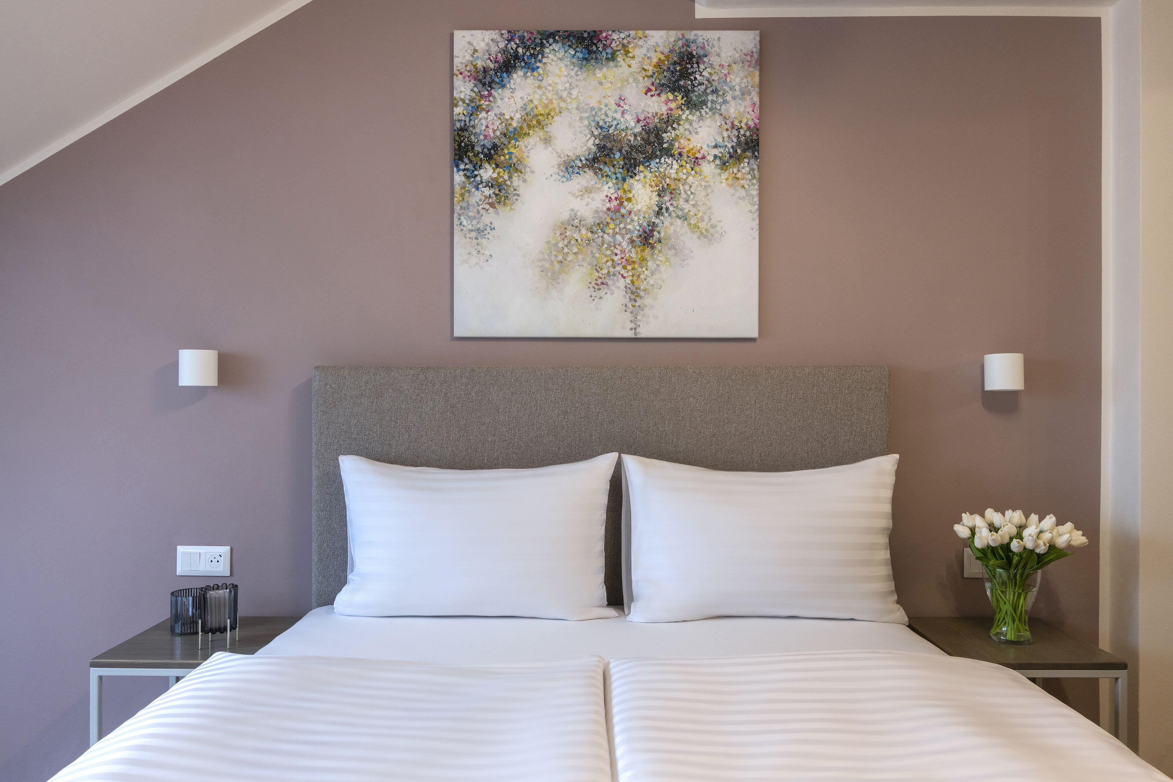Una cama bien tendida con sábanas blancas, cabecera gris y una obra de arte abstracta colgada en la pared.