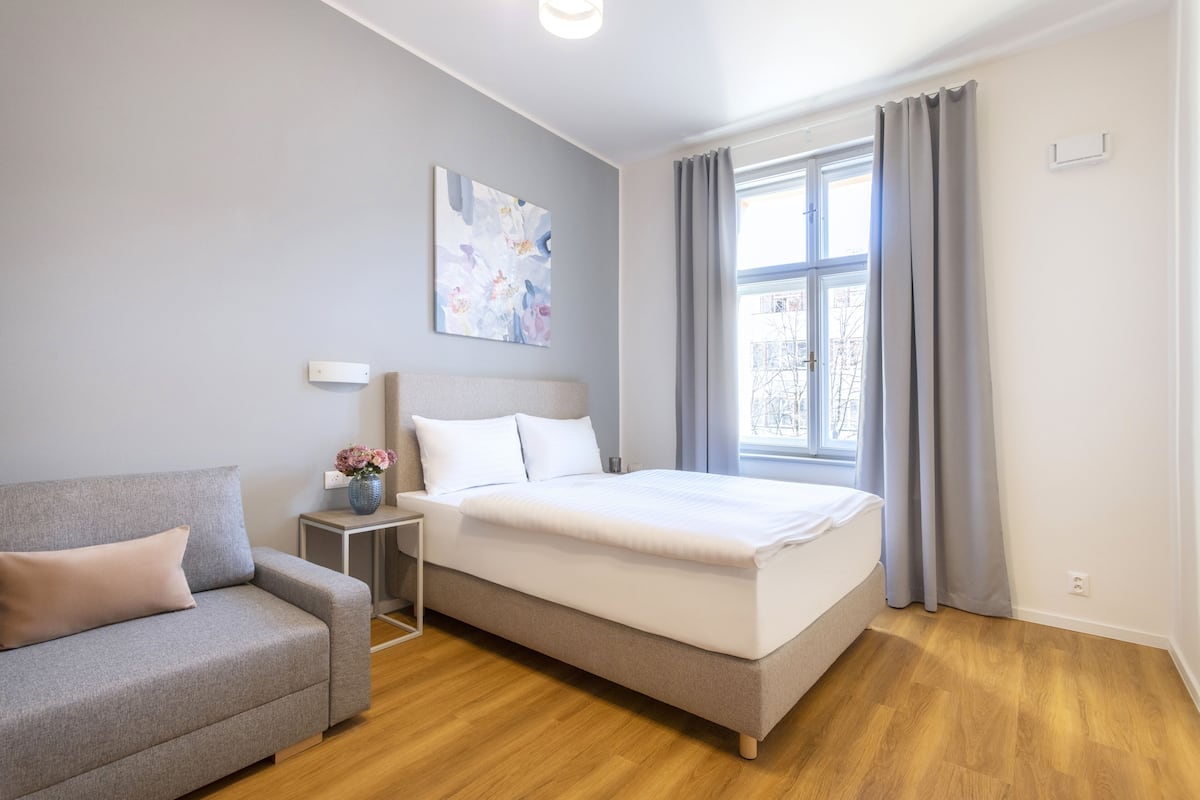 Un dormitorio moderno con cama, sofá, mesita de noche y ventana con cortinas.