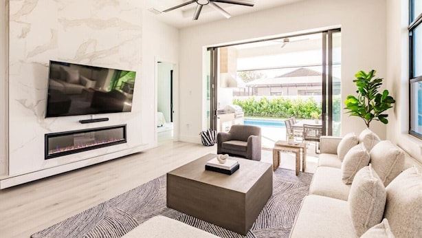 A modern living room with a flat-screen TV, a fireplace, a ceiling fan, and a view of a pool and greenery outside. - Naples Calming Waves Luxury Beach Home Heated Pool/Spa Walk 2 Beach (Naples)
