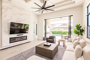 A modern living room with a flat-screen TV, a fireplace, a ceiling fan, and a view of a pool and greenery outside.
