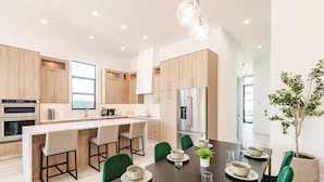 A modern kitchen with wooden cabinets, a central island with bar stools, a dining table set for four, and a refrigerator. - Naples Calming Waves Luxury Beach Home Heated Pool/Spa Walk 2 Beach (Naples)