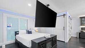 Ein modernes Hotelzimmer mit einem großen Bett, einem flachbildfernseher an der Wand, einem dunklen Holzschreibtisch und einer Glastür zum Badezimmer.