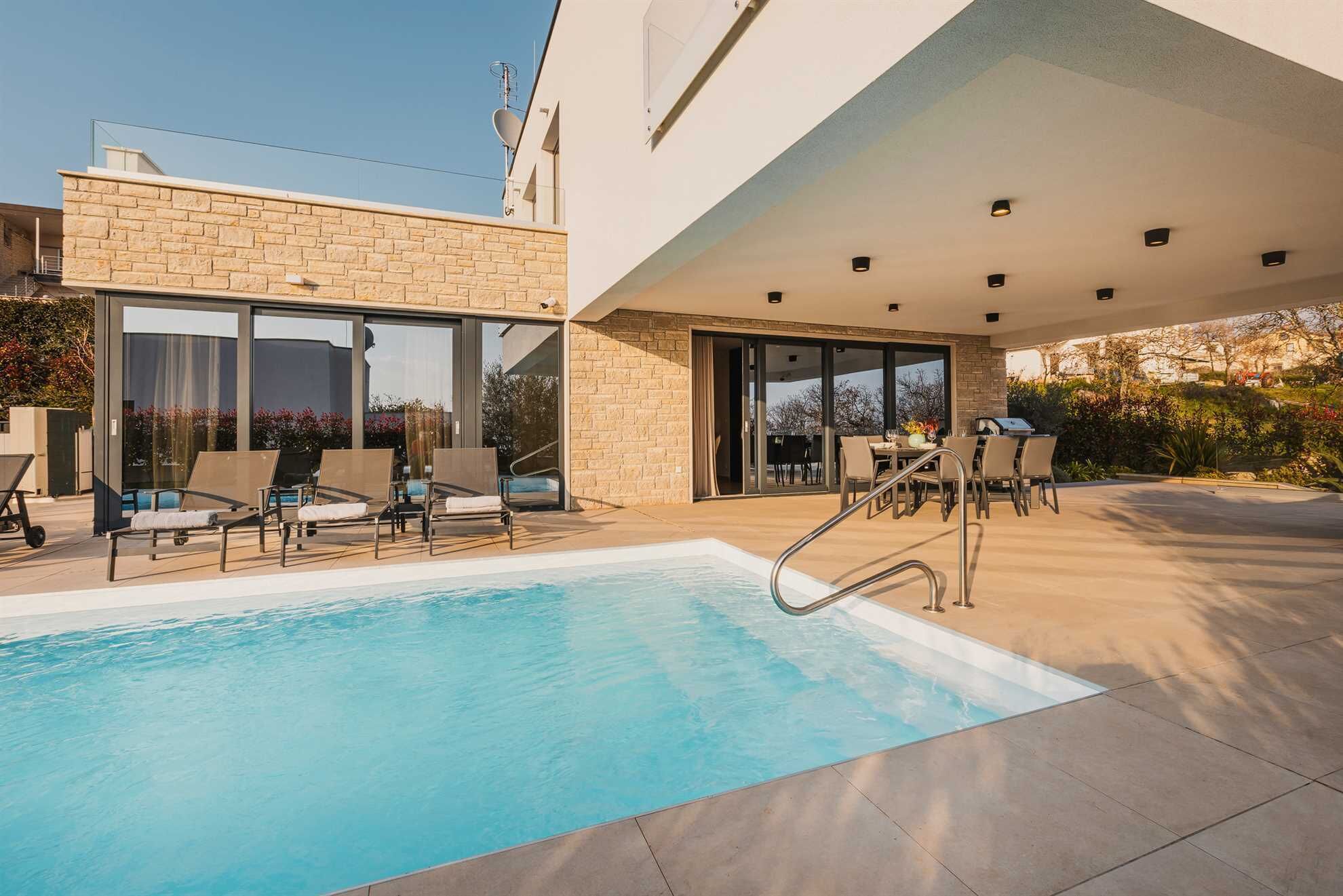 A modern outdoor pool area with lounge chairs and a barbecue setup.