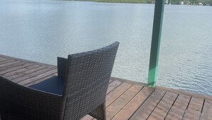 Una terraza de madera con una silla de mimbre y una mesa, con vista a un lago tranquilo y una orilla lejana.