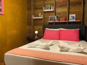 Ein ordentlich bezogenes Bett mit rosa Kissen und weißen Handtüchern, eine Nachttischlampe sowie Holzregale mit Büchern und Dekoration.