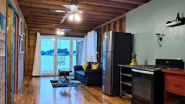 Una habitación amplia con pisos de madera, ventilador de techo, refrigerador, estufa y vista al exterior.